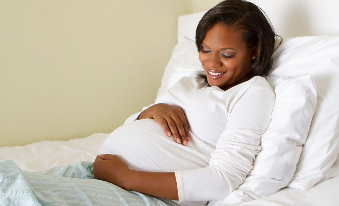 An African American woman who is pregnant smiling in her bed and holding her stomach
