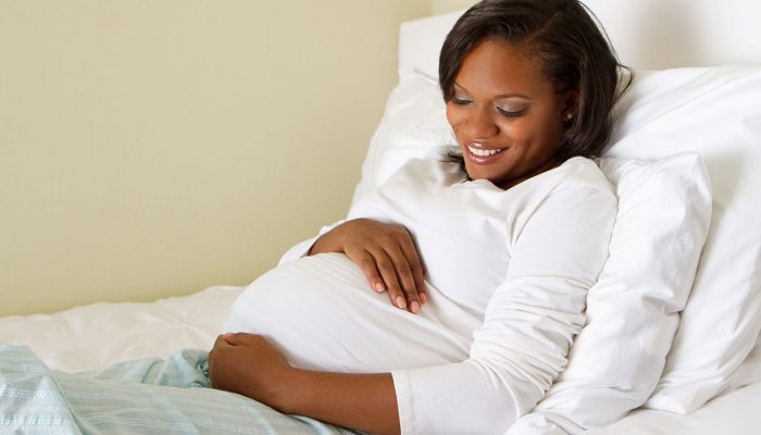 An African American woman who is pregnant smiling in her bed and holding her stomach