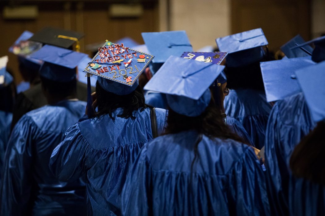 Graduates stand together in blue gowns, with blue caps on their heads. One cap reads “Oh I’m not tired yet, next, 2021.”