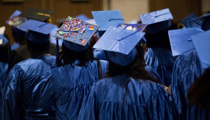 Graduates stand together in blue gowns, with blue caps on their heads. One cap reads “Oh I’m not tired yet, next, 2021.”
