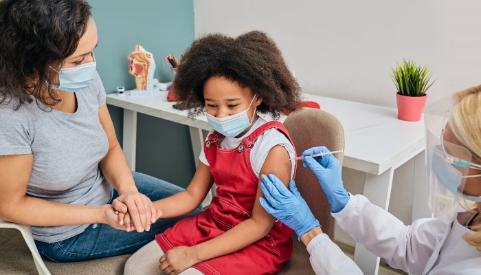 Child wearing a mask and holding her mother's hand while she receives a vaccine