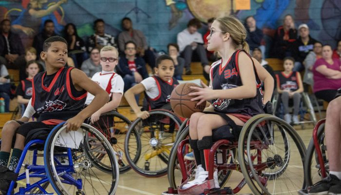 A girl in her wheelchair going to shoot the basketball while surrounded by teammates.