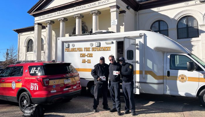 three men standing in front of large van with open door and marked fire department SUV with columned library behind them in daylight