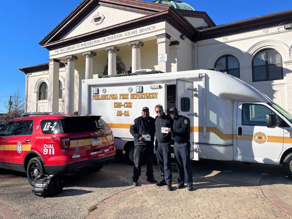 three men standing in front of large van with open door and marked fire department SUV with columned library behind them in daylight