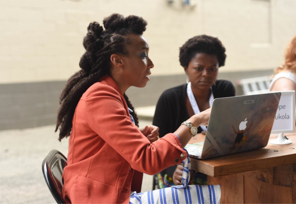 A woman is pointing to a laptop and talking to the woman sitting next to her.