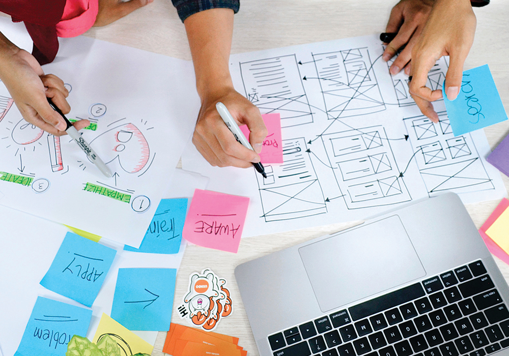 Hands are pointing and writing/drawing at a table covered in papers, sticky notes, and a laptop.