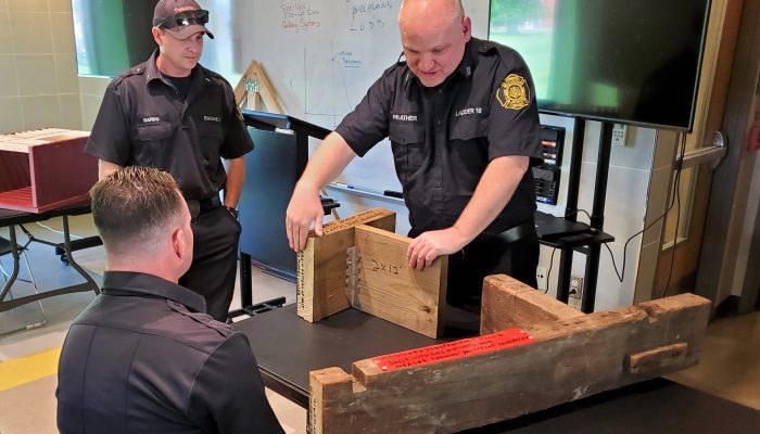 firefighter handles large pieces of wood on table in classroom as two other firefighters look on
