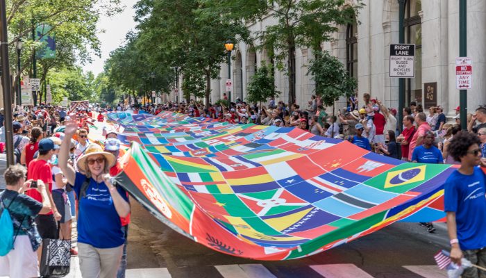 As pessoas estão andando por uma rua da Filadélfia segurando uma bandeira gigante colorida que apresenta imagens menores de bandeiras de diferentes países do mundo. Uma multidão de pessoas se alinha nos dois lados da rua.