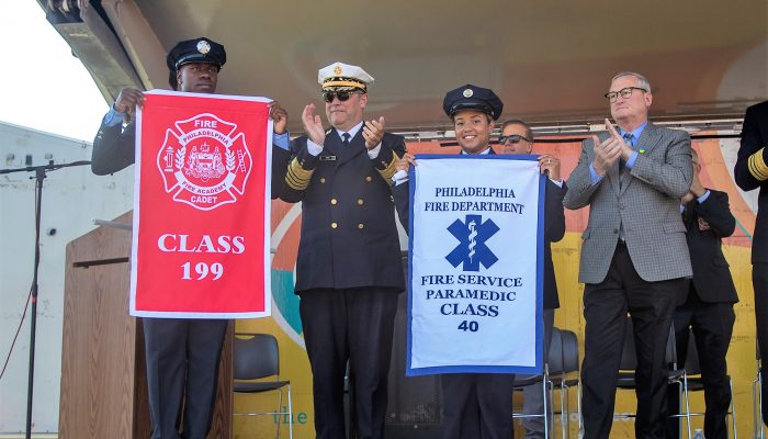 mayor and fire commissioner standing and clapping on stage next to firefighter and paramedic holding class banners at graduation ceremony