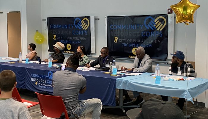 Six panelists sit behind tables covered with a Community Resource Corps table cloth.