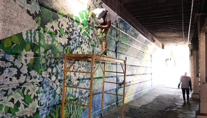 The Emerald Street Mural, composed of many triangular sections of paintings of green plants. The mural is still in progress. There is yellow scaffolding in front of the mural, and a silhouette of a person standing in the distance.