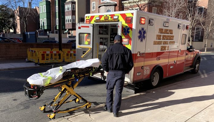 paramedic stands near empty stretcher at back of ambulance with bay doors open in daylight