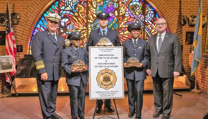 firefighters and paramedics in dress uniform stand with man in suit in front of stained glass window while holding trophies near poster for firefighter and ems providers of the year