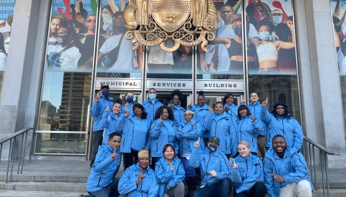 Membros do Community Resource Corps posam nos degraus em frente ao Edifício de Serviços Municipais. Eles levantam o dedo indicador para indicar que são a primeira coorte.