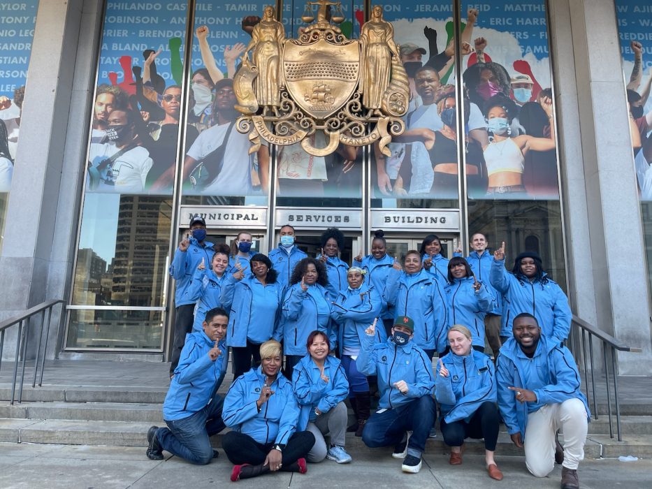 Community Resource Corps members pose on steps in front of the Municipal Services Building. They hold up their index finger to point that they're the first cohort.