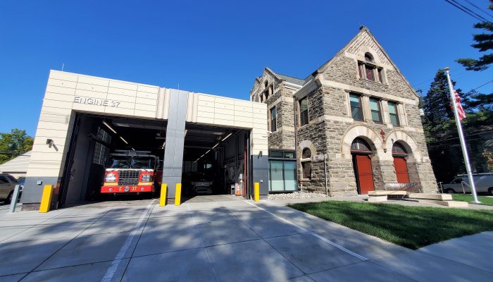 fire engine in modern garage next to historic three-story stone fire station with lawn and flag in daylight