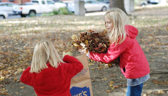 Two little girls put a pile of leaves in a tall brown bag in the park.