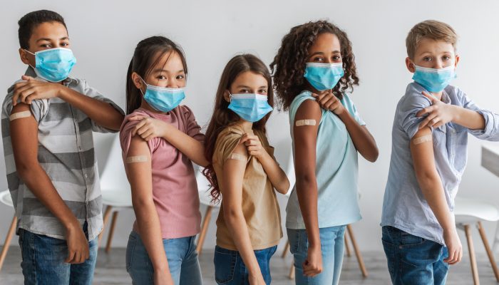Five children of different races and ethnicities wearing masks and holding up their sleeves to show bandages from vaccinations