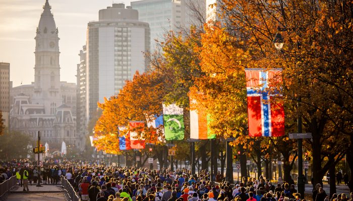 Thousands of runners line up along Benjamin Franklin Parkway and begin their Marathon journey racing towards City Hall. The street is lined with fall colored trees and flags of many different nations.