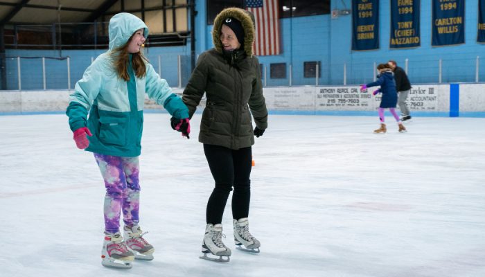 Two girls holding hands while ice skating in an indoor ice rink.