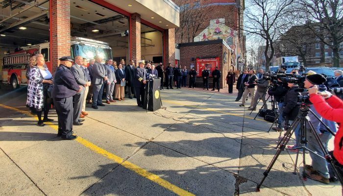 outdoor press conference at firehouse with TV cameras and media on right side and officials standing in driveway in front of fire engine on left side in daylight