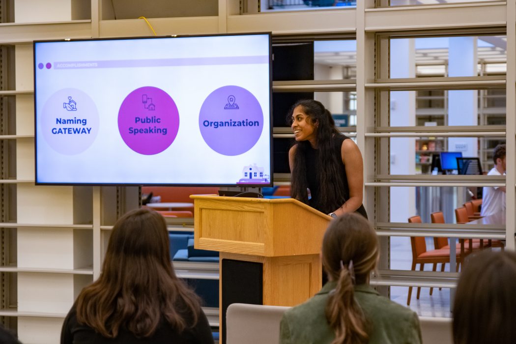 A Serve VISTA speaks in front of a crowd from behind a podium. Behind them is a large screen showing a slide from a presentation. On the slide there are three circles with text inside that reads, "Naming GATEWAY, Public Speaking, Organization."