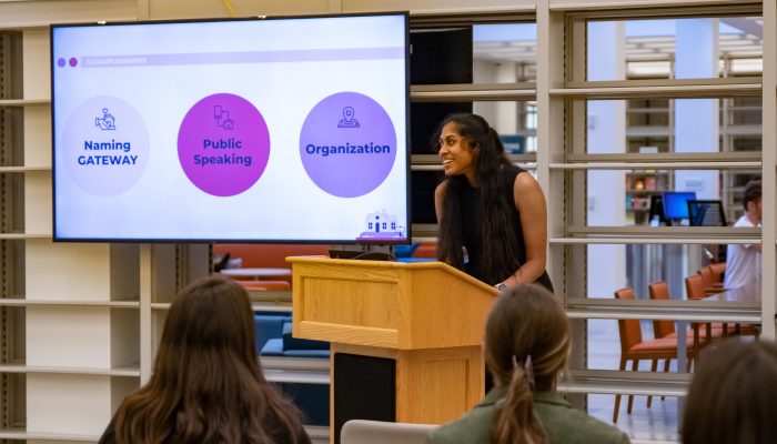 A Serve VISTA speaks in front of a crowd from behind a podium. Behind them is a large screen showing a slide from a presentation. On the slide there are three circles with text inside that reads, "Naming GATEWAY, Public Speaking, Organization."