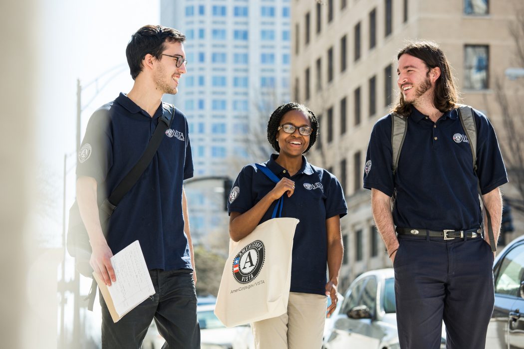 Three VISTA members wearing branded navy blue polos walk down a city street while talking.