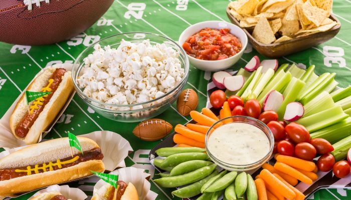 Healthy snacks, like cut vegetables and popcorn, sitting on a green tablecloth with a football