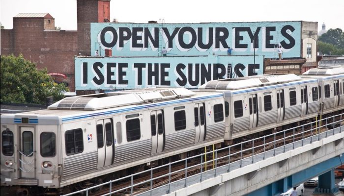 a septa market frankford line train passes in front of a mural on the side of a building that says open your eyes I see the sunrise