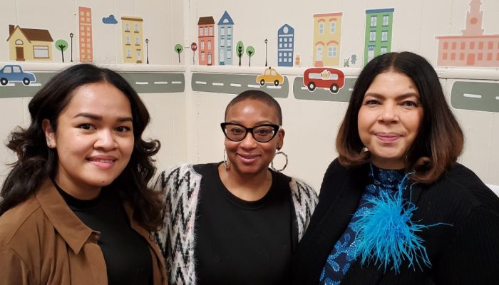 Three women pose for a picture at a learning and play area in a homeless shelter.