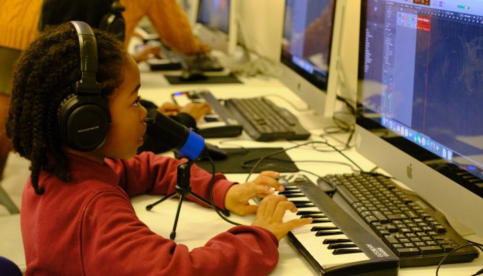 A boy sings into a microphone and plays a keyboard while sitting at a computer in Music lab.