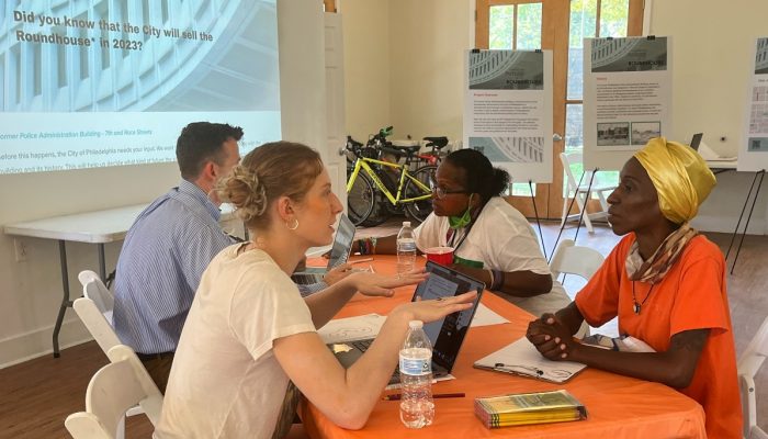 Four people sit at a folding table. They are having conversations with each other across laptops. In the background a power point projection says "did you know the city will sell the round house in 2023"