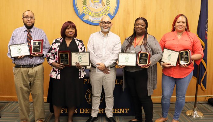 two men and three women standing, smiling and holding various plaques and awards with philadelphia fire department seal on wall behind them