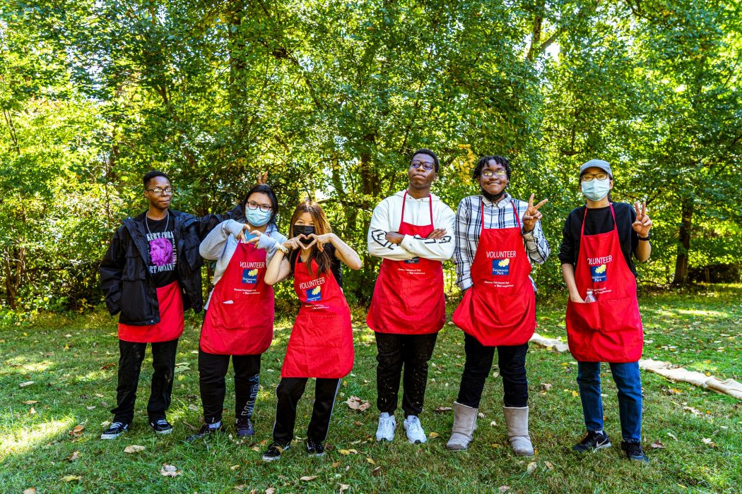 Six volunteers standing in front of a forest wearing red Fairmount Park Volunteers aprons.