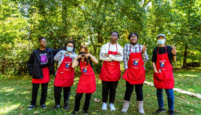 Six volunteers standing in front of a forest wearing red Fairmount Park Volunteers aprons.