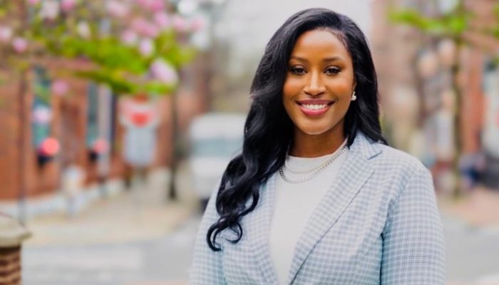 Tracie Johnson smiles at the camera. She's wearing a light blue checked blazer over a white shirt, standing on a street with flowering trees in the background.