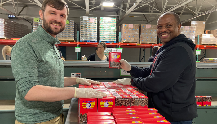 Two Philabundance volunteers look at the camera while arranging boxes of raisins for packing. One volunteer holds a box of raisins up. There are more volunteers in the background along an assembly line.