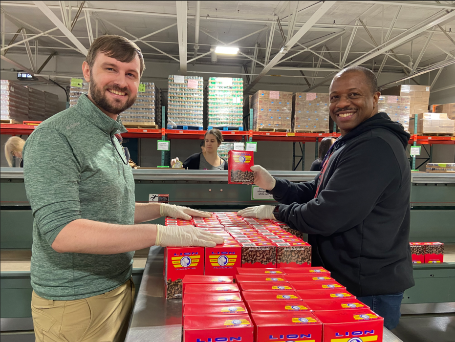 Two Philabundance volunteers look at the camera while arranging boxes of raisins for packing. One volunteer holds a box of raisins up. There are more volunteers in the background along an assembly line.