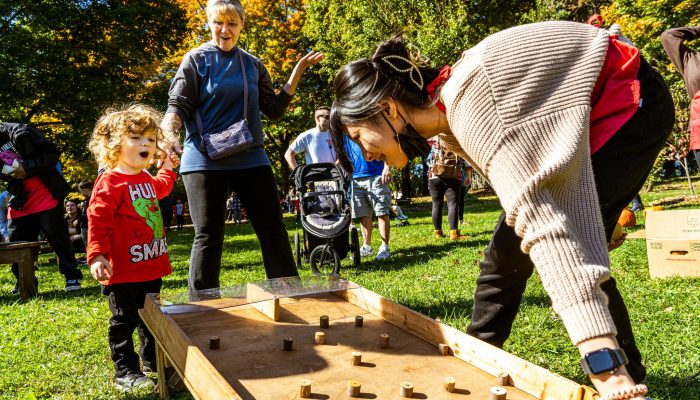 A toddler exclaims, looking at something a volunteer is setting up. The toddler is holding an adult's hand; the adult is standing further in the back. The volunteer is closer to the front and is preparing to lift a wooden set up for an activity.