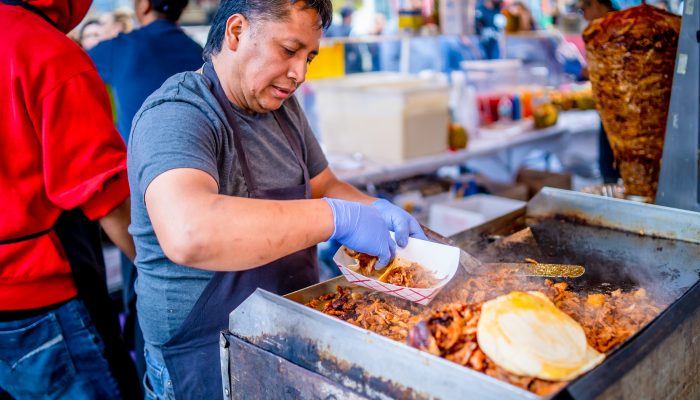 A man in an apron and blue latex gloves cooks over a flat-top grill. In his hand is a to-go basket that he is placing tacos into.In the background you can see other employees as well as the crowd waiting for their food.