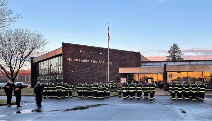 dozens of firefighter cadets standing in formation in bunker gear at dawn outside brick building labeled philadelphia fire academy