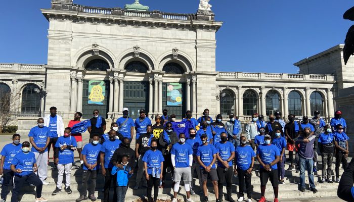 A group photo of Brothas Stroll attendees wearing blue event tee shirts from previous stroll. Standing in front of the Please Touch Museum