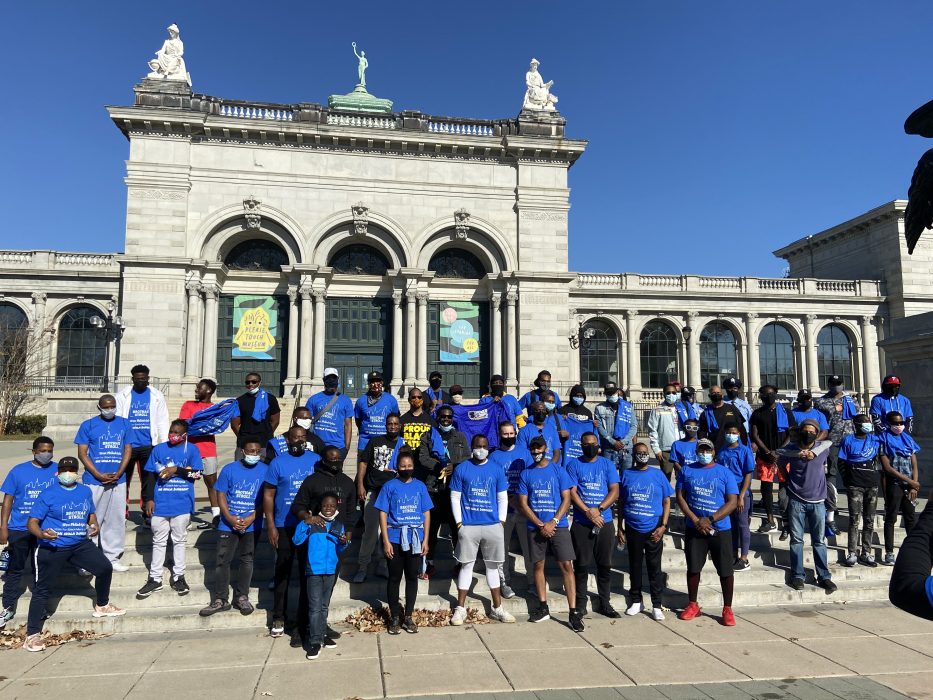 A group photo of Brothas Stroll attendees wearing blue event tee shirts from previous stroll. Standing in front of the Please Touch Museum
