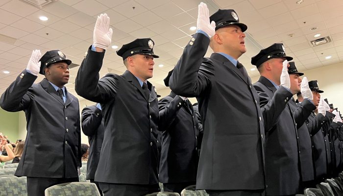 firefighter cadets in dress uniforms holding gloved hands up as they take the oath of office in an auditorium