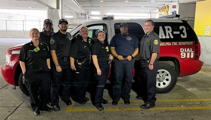 paramédicos uniformizados em pé com especialistas em saúde comportamental em frente ao SUV marcado com AR-3 na janela lateral traseira da garagem