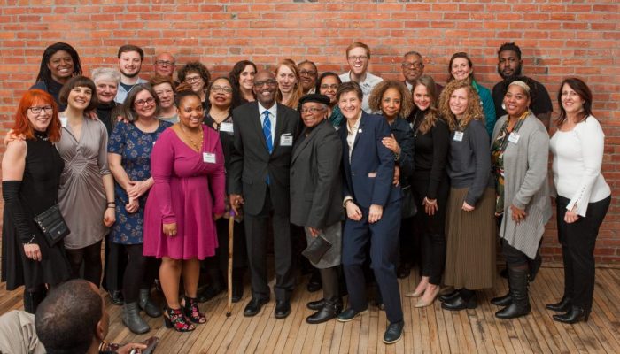 A group of people standing in front of a brick wall, they are posed for the camera and smiling.