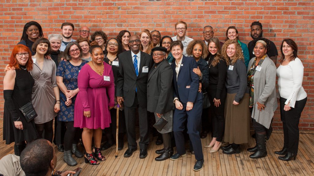 A group of people standing in front of a brick wall, they are posed for the camera and smiling.