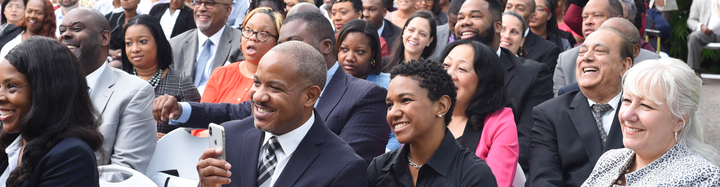 A large crowd listens and smiles as a presentation takes place.