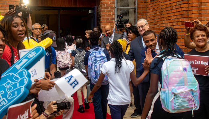 Mayor Kenney and Superintendent Watlington high fiving students as they walk into a school on the first day of school last year. There is also a crowd around them with foam fingers and welcome back signs